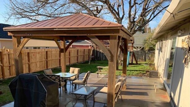 Une pergola en bois avec un toit en métal, un espace extérieur aménagé avec des tables et des chaises, et un barbecue.