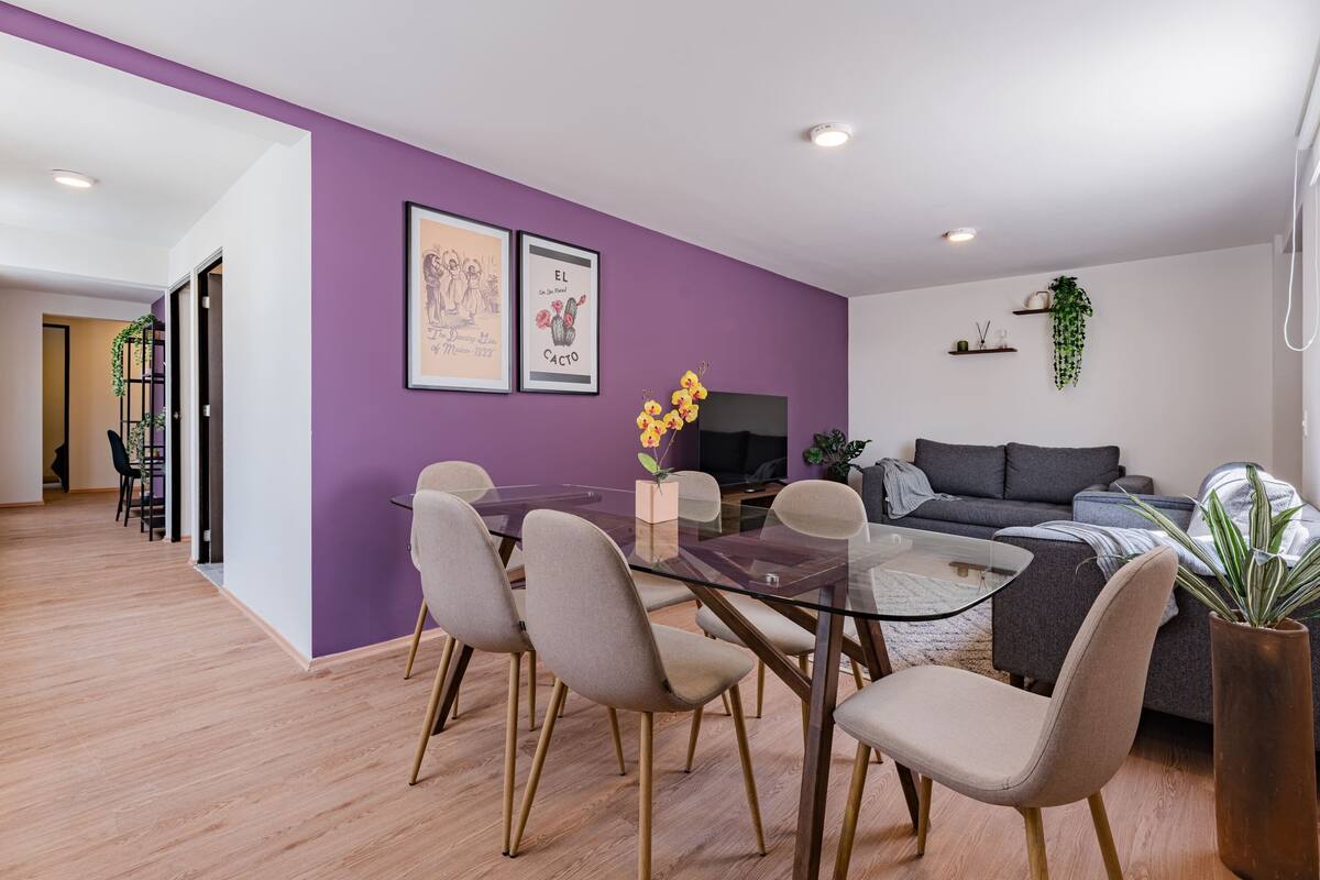 Una sala de estar moderna con una pared de acento púrpura, una mesa de comedor de vidrio con sillas, un sofá gris y un ventanal grande.