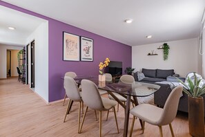 Un salon moderne avec un mur d’accent violet, une table à manger en verre avec des chaises, un canapé gris et une grande fenêtre.