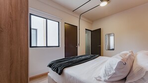 Un dormitori amb una finestra gran, un llit amb roba de llit blanca, una porta fosca i un mirall a la paret.