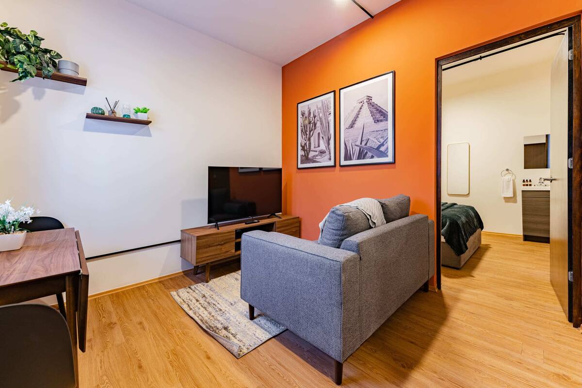 Una sala moderna con un sofá gris, piso de madera y un televisor sobre un mueble de madera. Hay dos cuadros en la pared naranja, una pequeña mesa con una planta, y una puerta que da al baño.