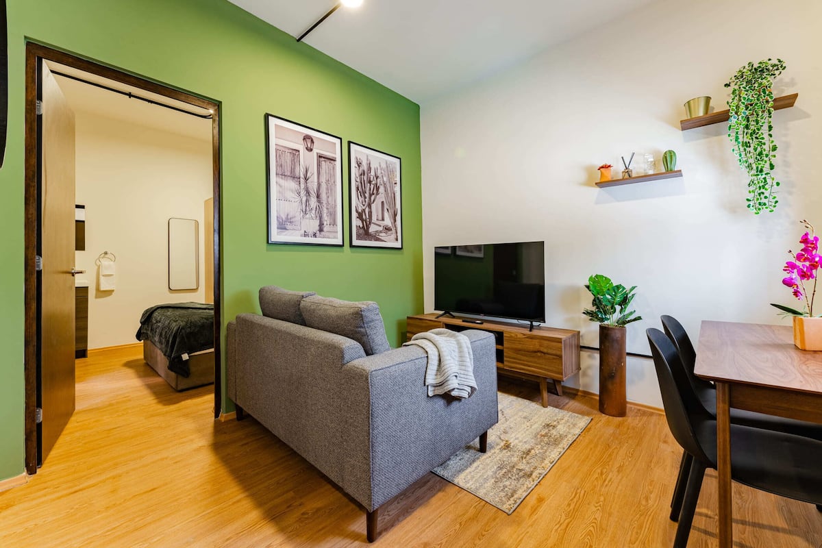 Una sala de estar moderna con un sofá gris, piso de madera, paredes verdes, un televisor de pantalla plana, una mesa de comedor con sillas y plantas decorativas.