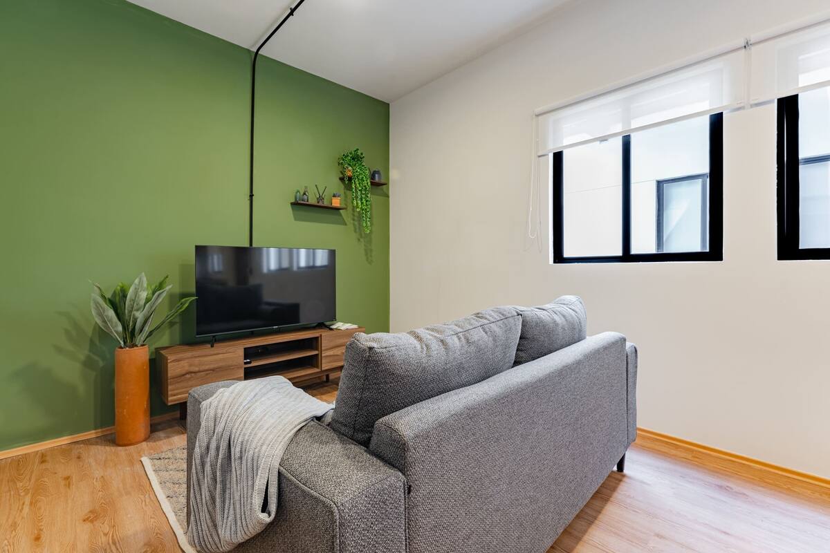 Una sala de estar con un sofá gris, un televisor sobre un mueble de madera, una pared verde y una ventana con persianas.