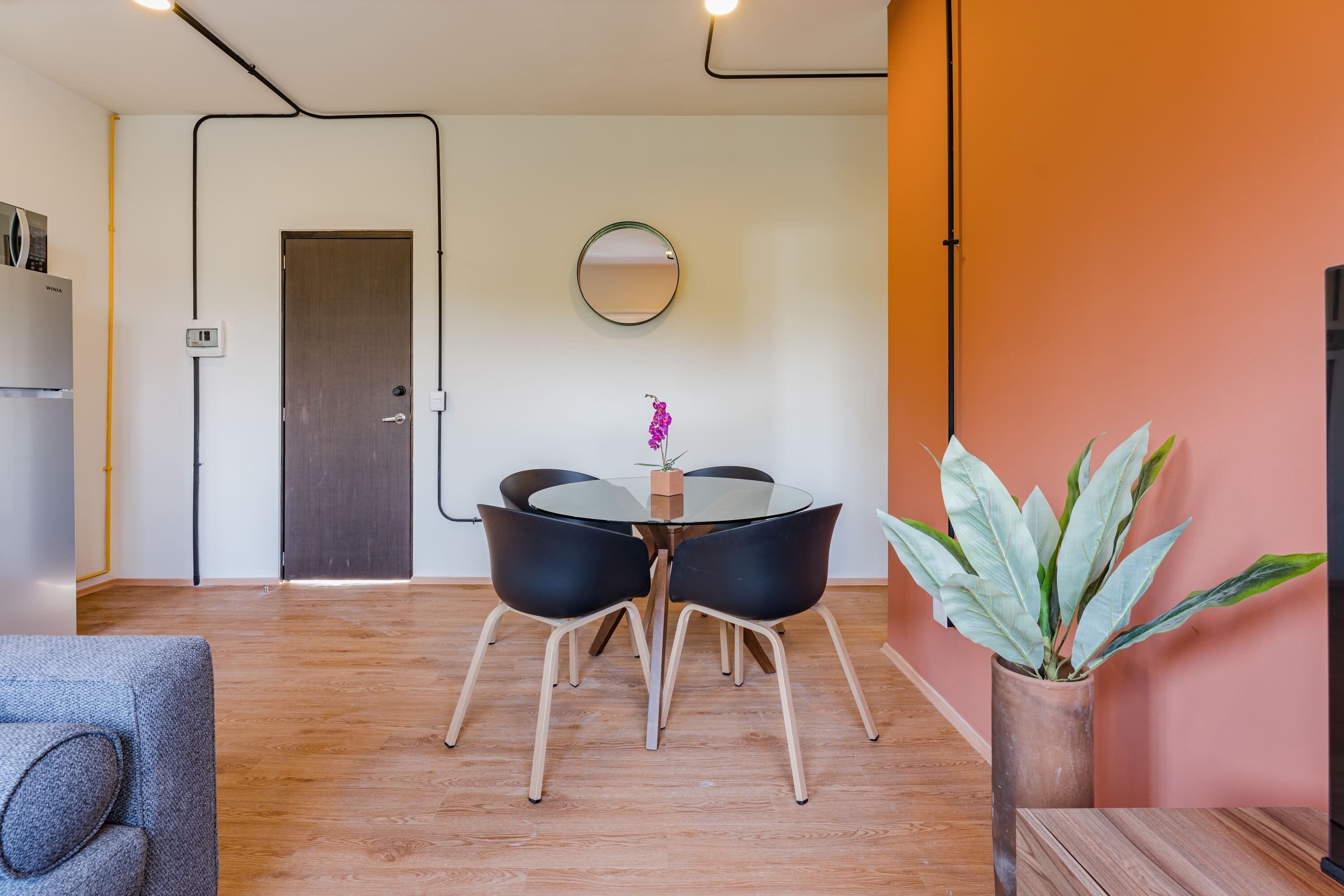 A modern living room with a glass dining table, black chairs, a wooden floor, a grey sofa, a potted plant, and a mirror on the wall.