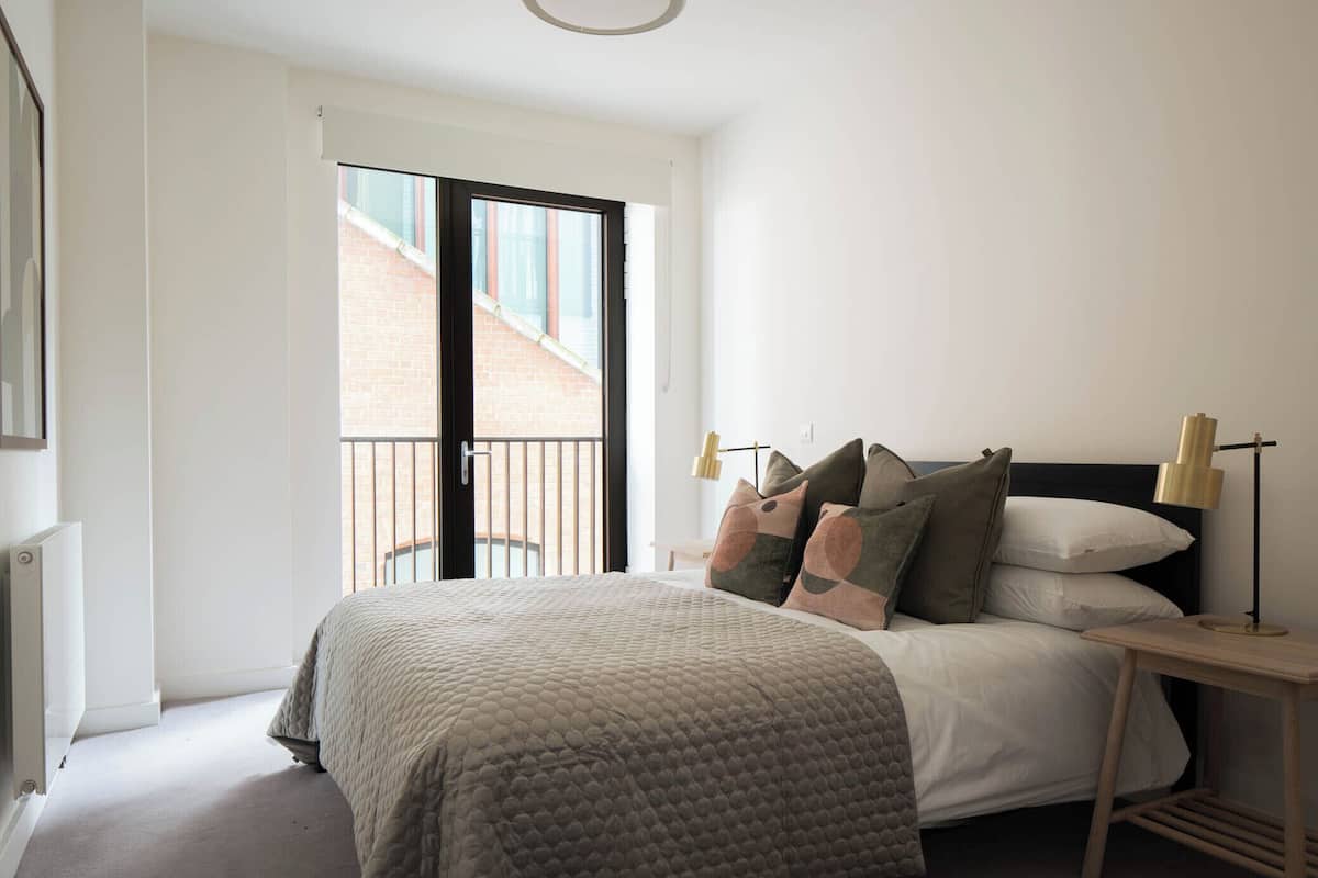 A bedroom with a large bed, a nightstand with a lamp, and a view of a brick building through a sliding door.