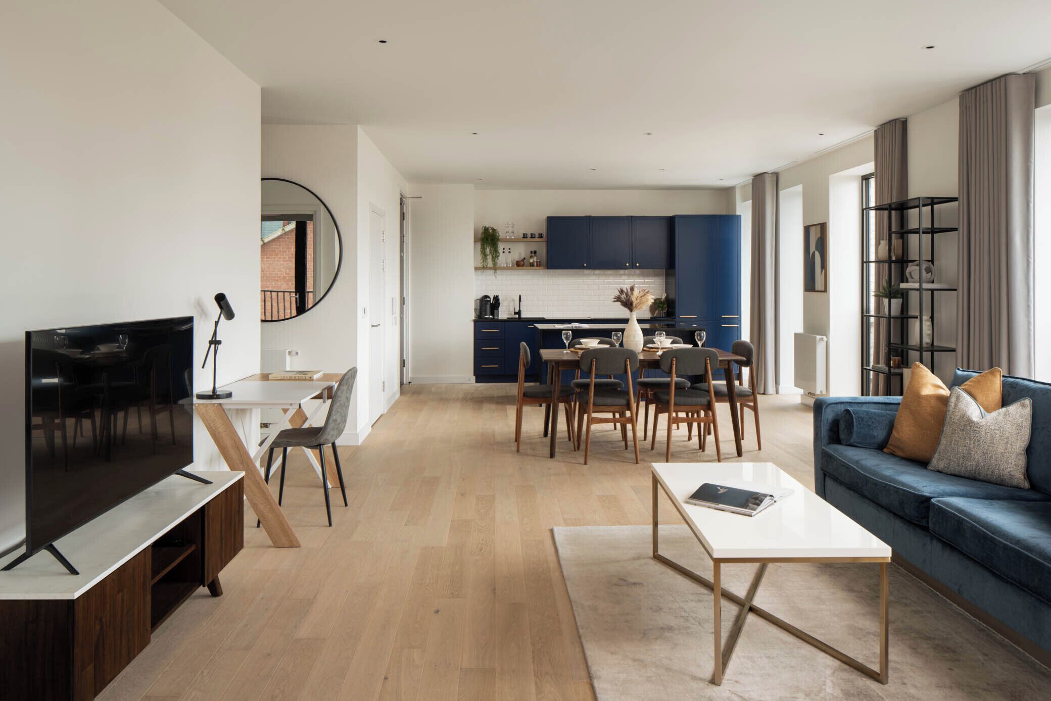 A modern living room with a flat-screen TV, a dining area with a table and chairs, a sofa with cushions, and a kitchen area with blue cabinets and a white countertop.