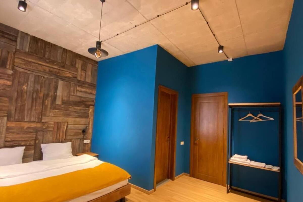 A bedroom with a bed, wooden wall paneling, a blue wall, a wooden door, a wooden bench, and a towel rack with hangers.