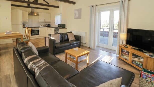 Ein modernes Wohnzimmer mit einer schwarzen Ledersofa, einem Holztisch und einem Fernseher auf einem Holzbodenständer. Im Hintergrund ist eine Küche mit weißen Schränken und einer Mikrowelle zu sehen.