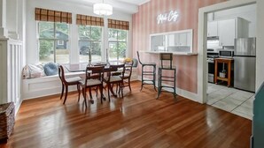 Un coin repas avec une table et des chaises en bois, un banc près de la fenêtre et une cuisine visible à travers une porte ouverte.