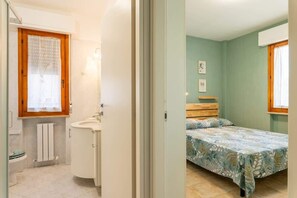 Quarto com cama, janela e banheiro com pia e vaso sanitário.
