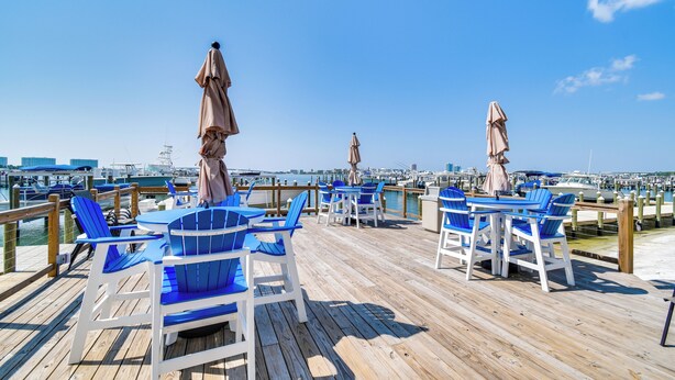 Eine Holzterrasse mit blauen Stühlen und Tischen, Blick auf die Marina mit Booten und einem strahlend blauen Himmel.