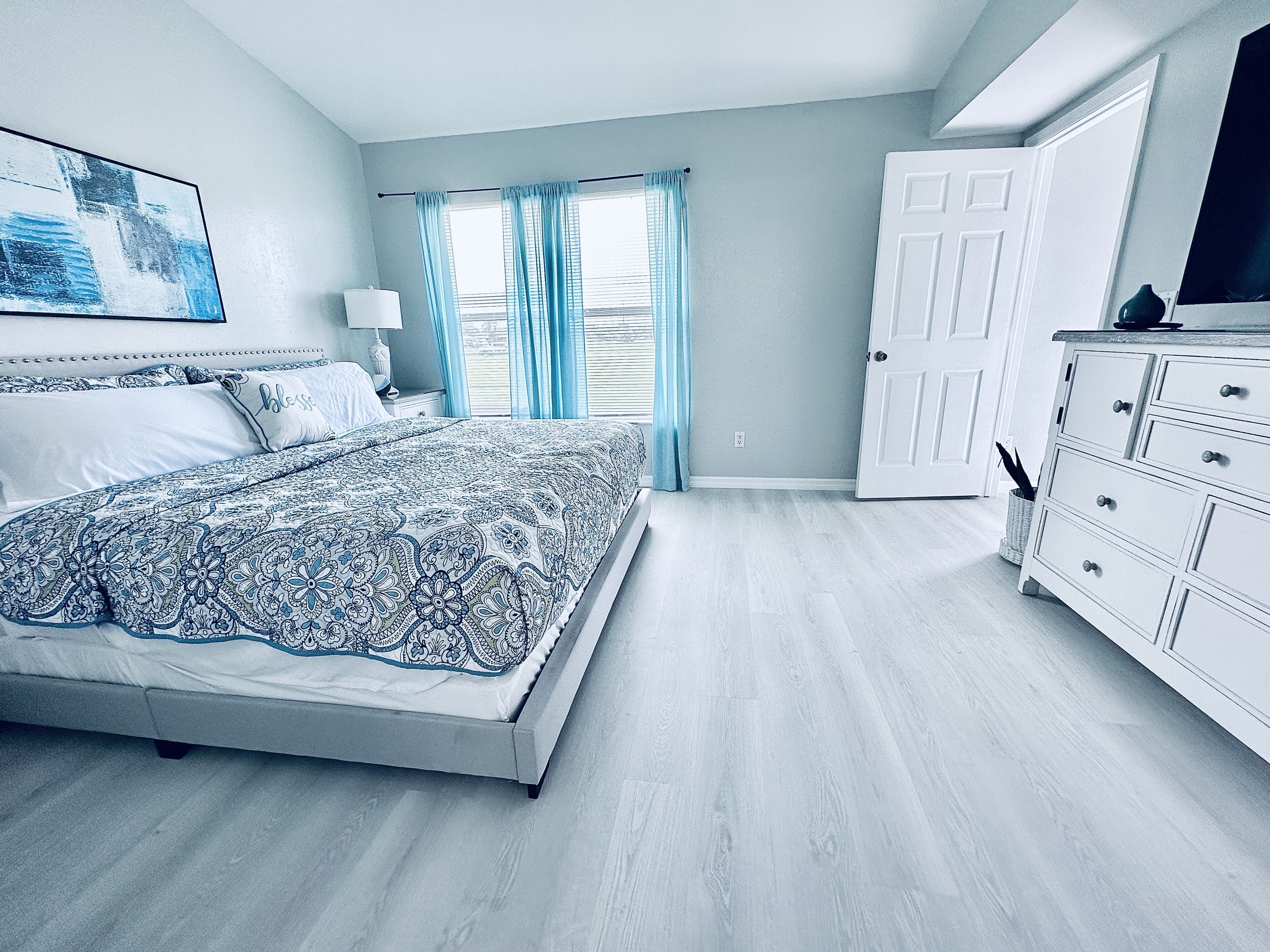 Une chambre à coucher avec un lit, une commode, une table de chevet, une lampe et une fenêtre avec des rideaux.