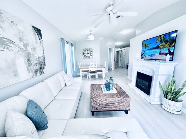 A modern living room with a white sofa, a coffee table, a fireplace, and a TV on the wall.
