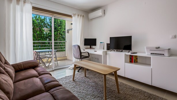 Una sala de estar moderna con un sofá marrón, una mesa de madera, un escritorio con dos monitores, un televisor y una impresora. Hay un balcón con una mesa y sillas, y un equipo de aire acondicionado blanco en la pared.