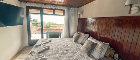 Un dormitorio con techo de madera, una cama grande con colcha, una mesita de noche con lámpara y vistas a través de un ventanal.