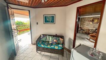 Una habitación con sofá de mimbre, una pequeña mesa, un cuadro en la pared y vistas al exterior.