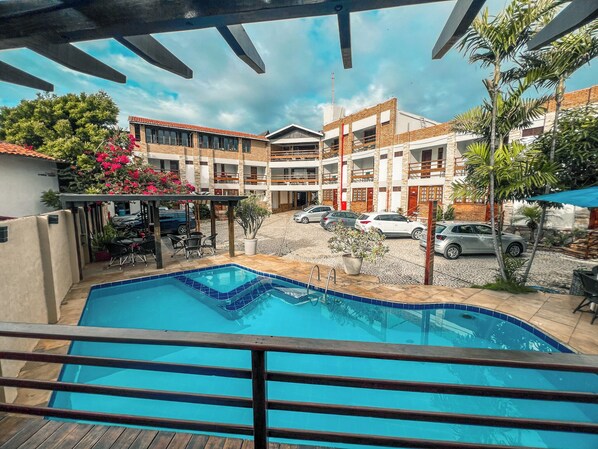 Une piscine avec une terrasse en bois au premier plan, entourée d’un immeuble à plusieurs étages doté de balcons et d’un parking où sont garées des voitures.