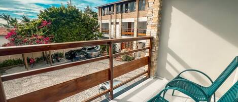 Un balcón con baranda de madera, dos sillas verdes y vistas a un edificio con varios balcones y coches aparcados.