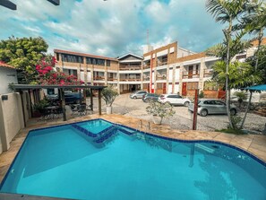 Une piscine dotée d’une terrasse carrelée, entourée d’un immeuble à plusieurs étages avec des balcons et d’un parking où sont stationnées des voitures.