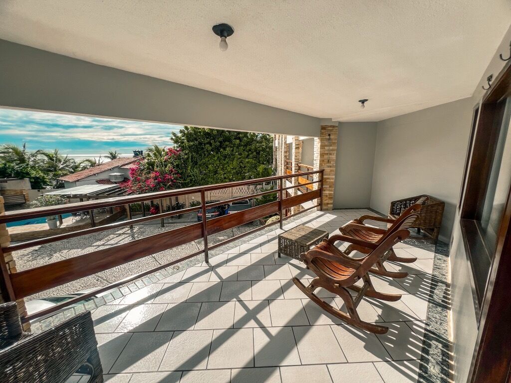 A balcony with a view of a pool and greenery, wooden rocking chairs, and a tiled floor.