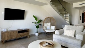 Una sala moderna con una televisión de pantalla plana, un mueble de madera, una mesa redonda de centro, una silla de mimbre y una escalera.