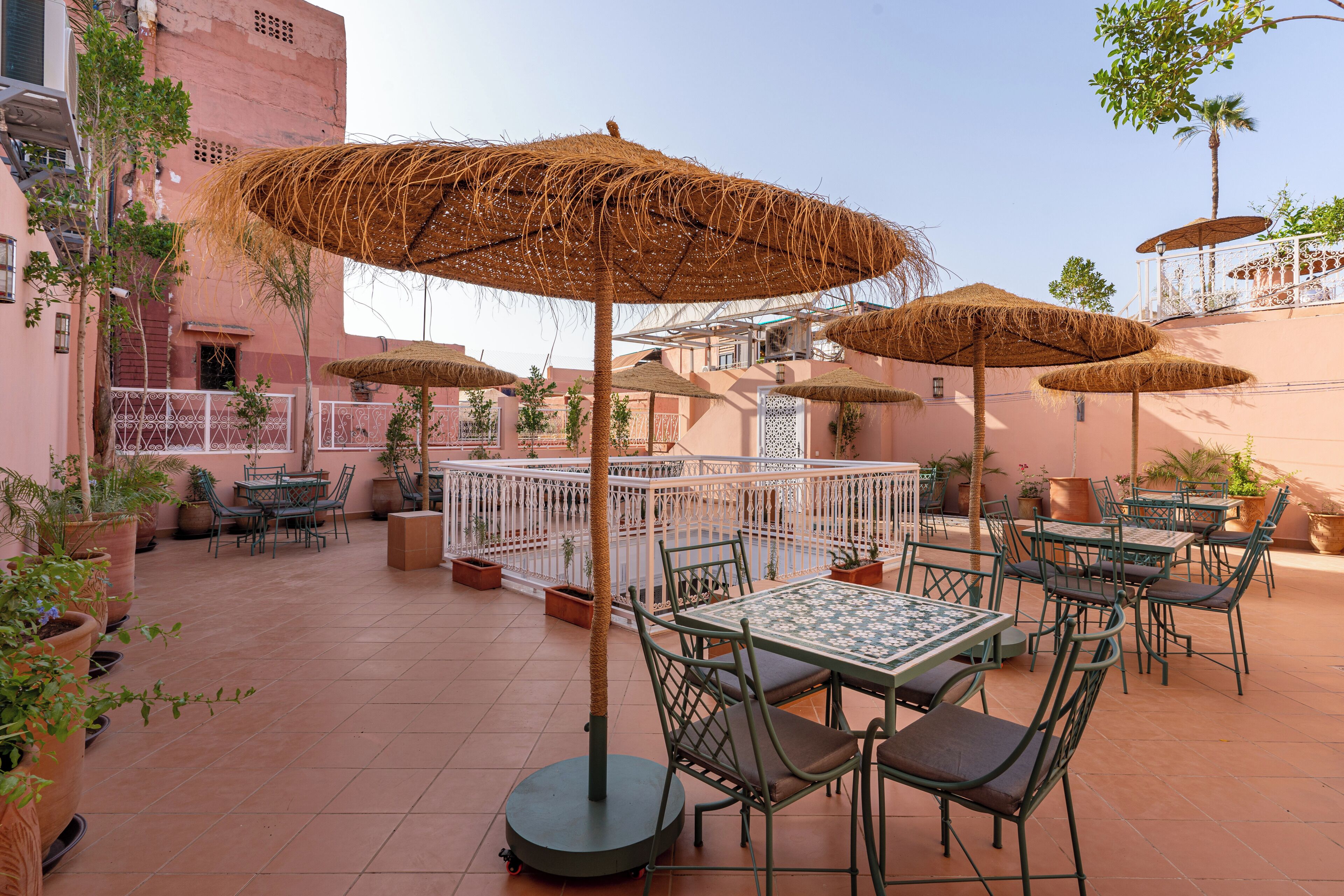 Une terrasse extérieure avec des tables et des chaises, entourée de plantes en pot et ombragée par des parasols en chaume.