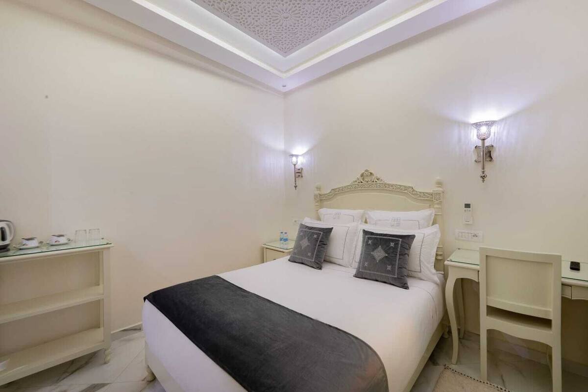 A hotel room with a bed, bedside tables, a nightstand, a desk, and a shelf with items.