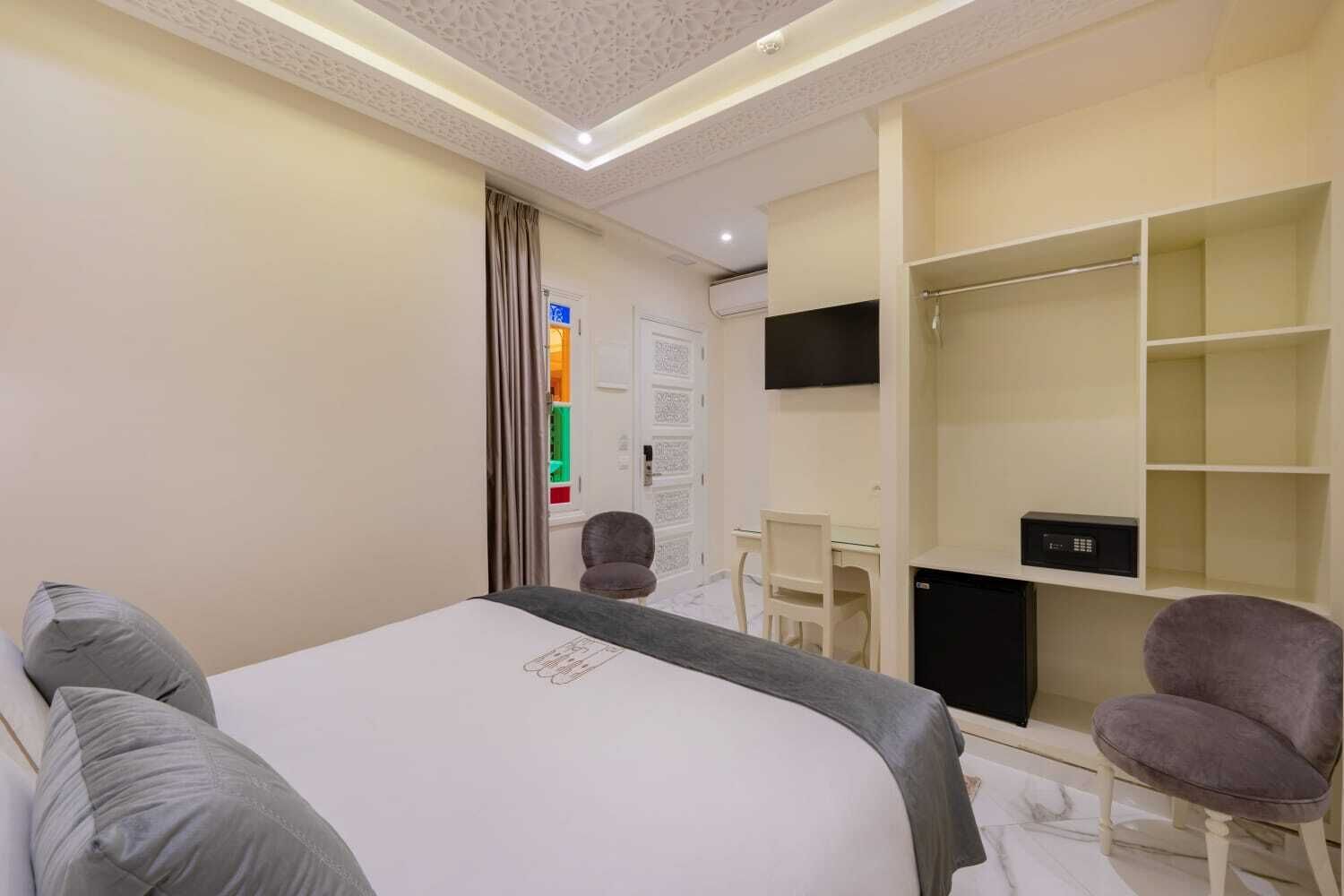 A hotel room with a bed, two chairs, a desk, a TV, and a wardrobe.