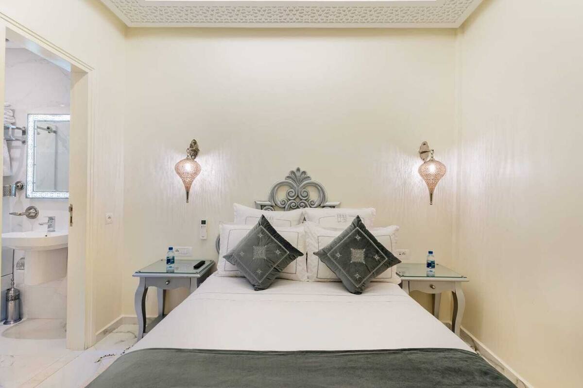 A bedroom with a bed, two bedside tables, a nightstand, a mirror, and two wall-mounted lamps.