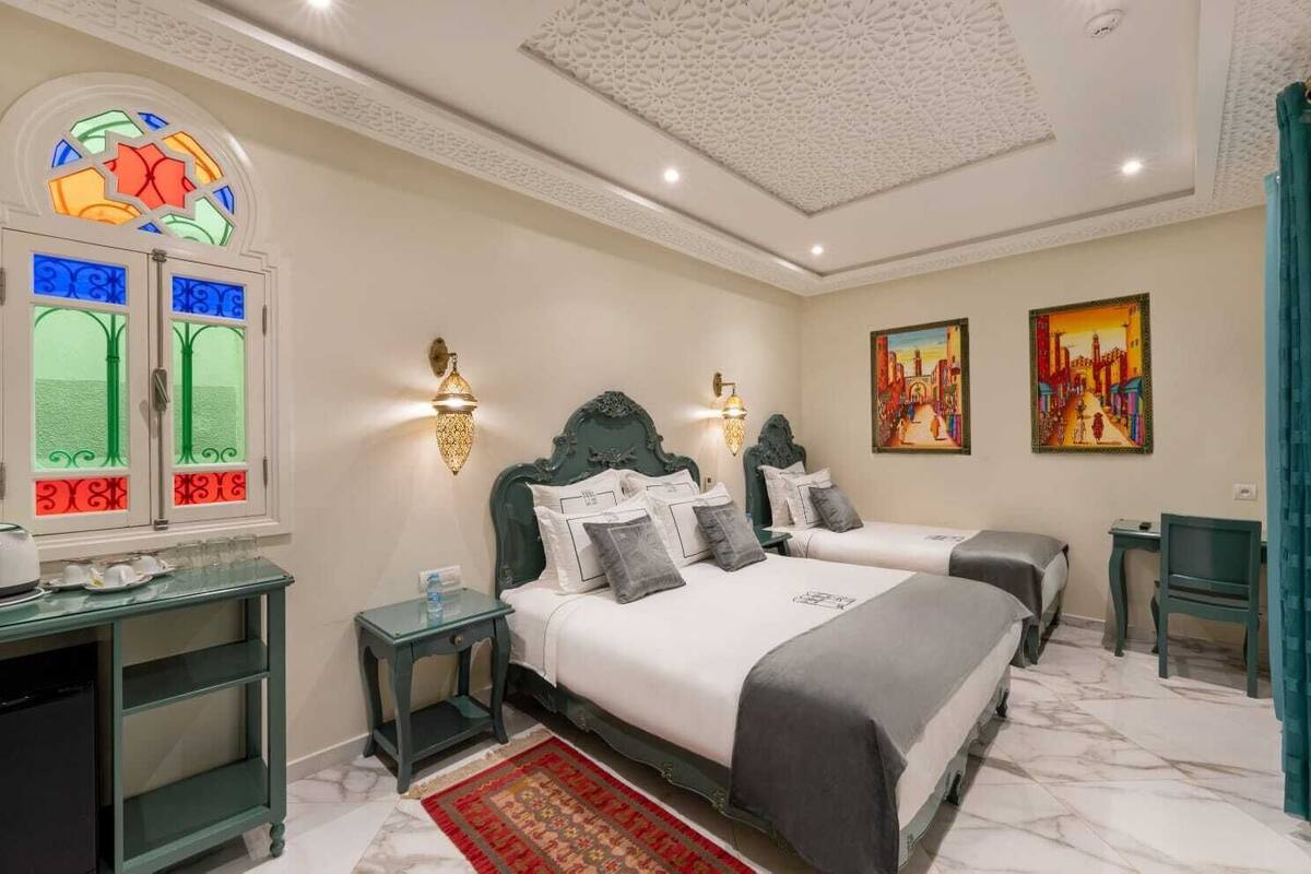 A hotel room with two beds, a glass table with cups and saucers, a small side table, and a decorative window.