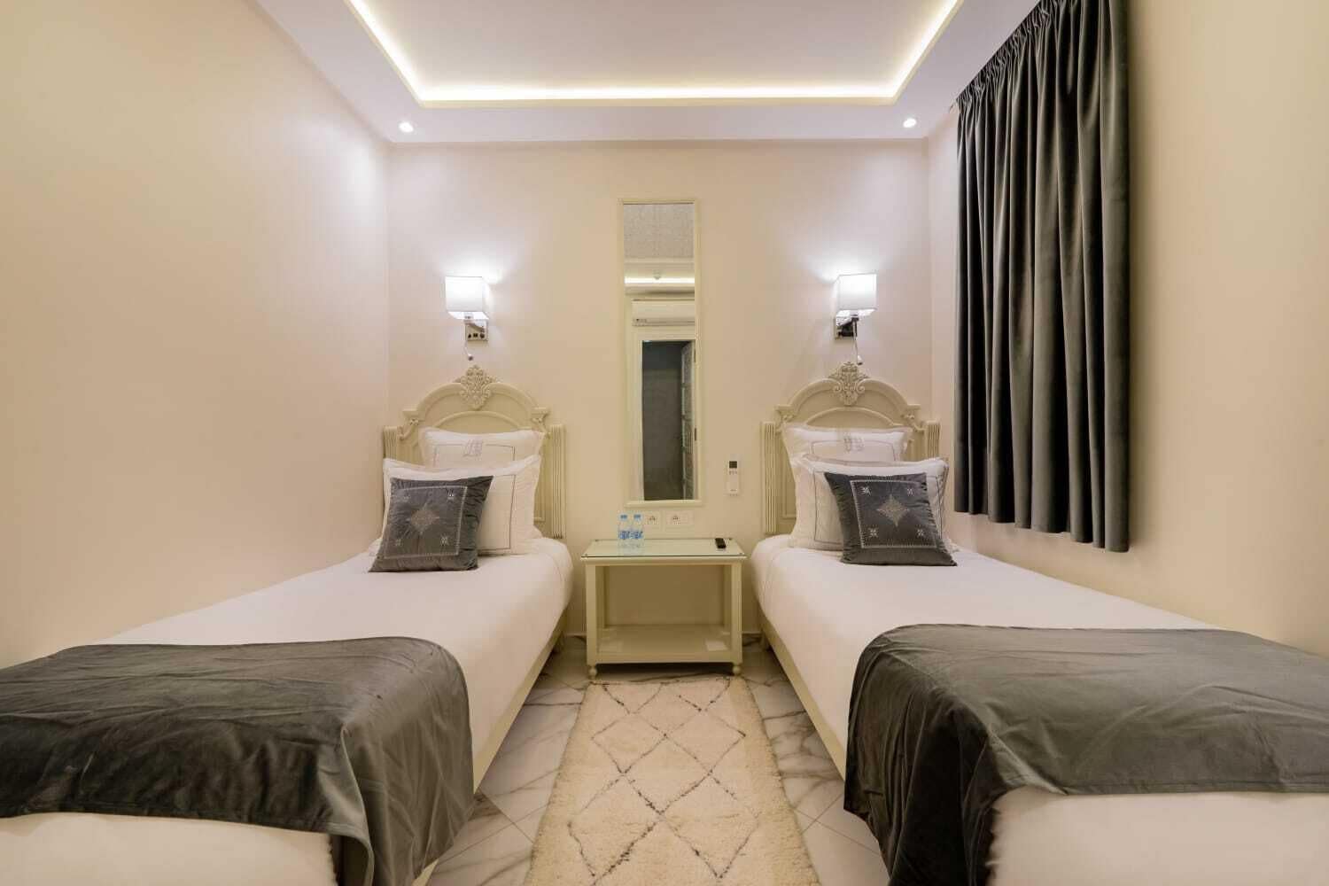 A hotel room with two single beds, a small bedside table, a mirror, and a window with curtains.