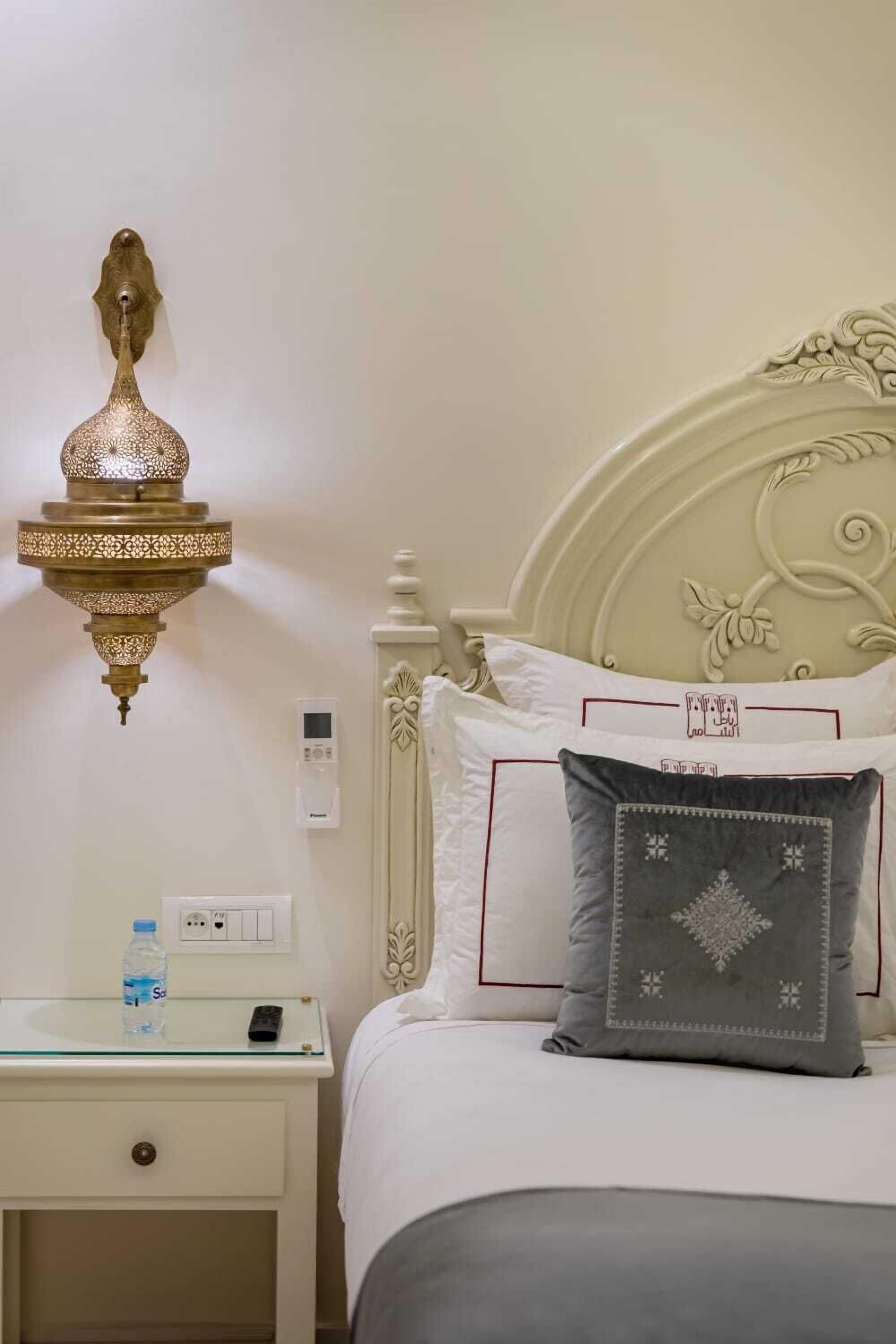 A neatly made bed with decorative pillows, a bedside table with a water bottle and remote control, and a wall-mounted light fixture.