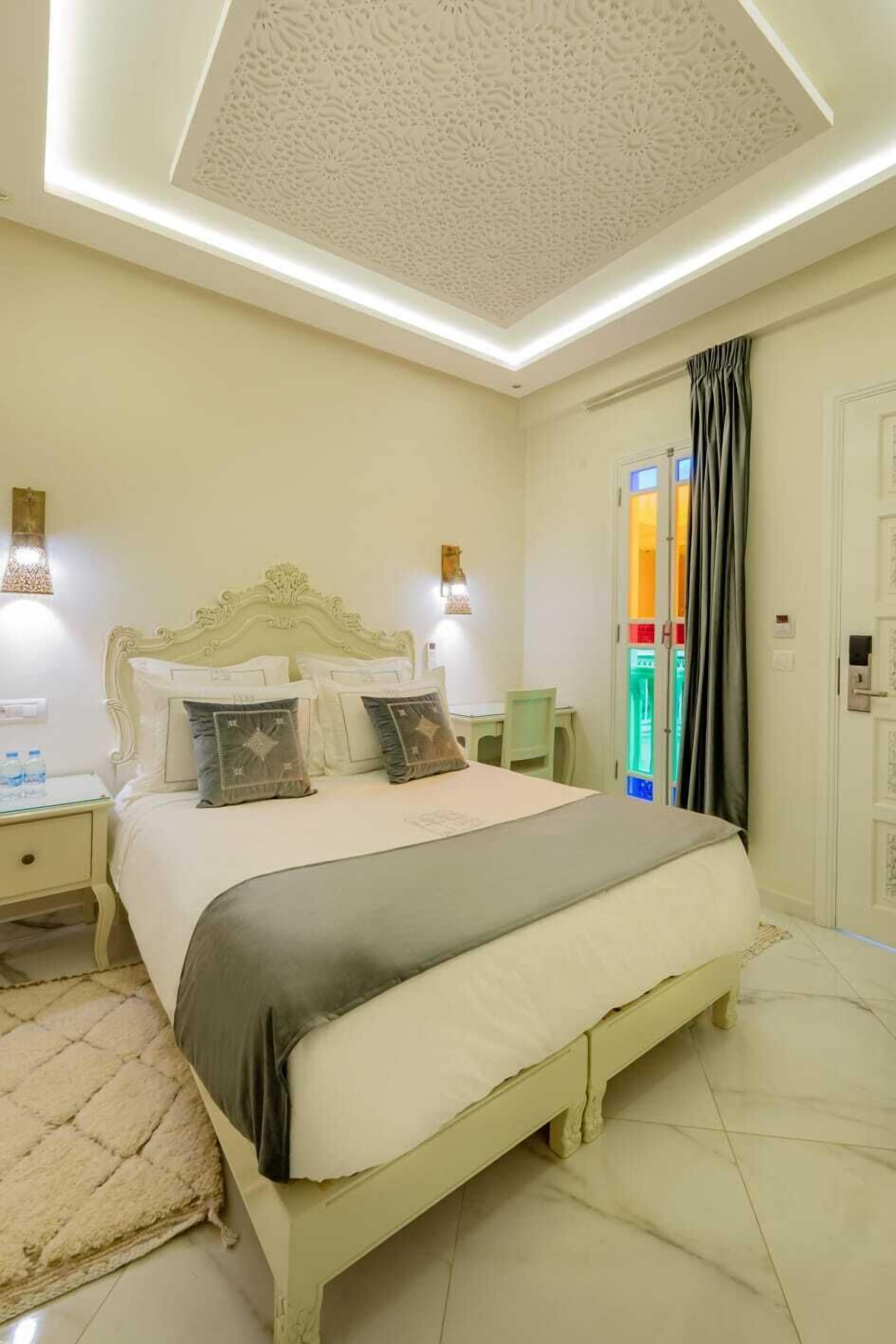 A bedroom with a bed, nightstands, a wall-mounted lamp, and a door with a colorful glass panel.