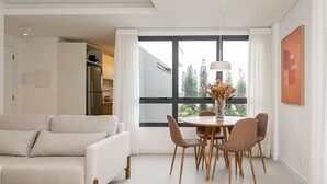 Uma sala de estar moderna com um sofá branco, mesa de jantar com cadeiras, um vaso com flores secas e uma grande janela com vista para árvores.