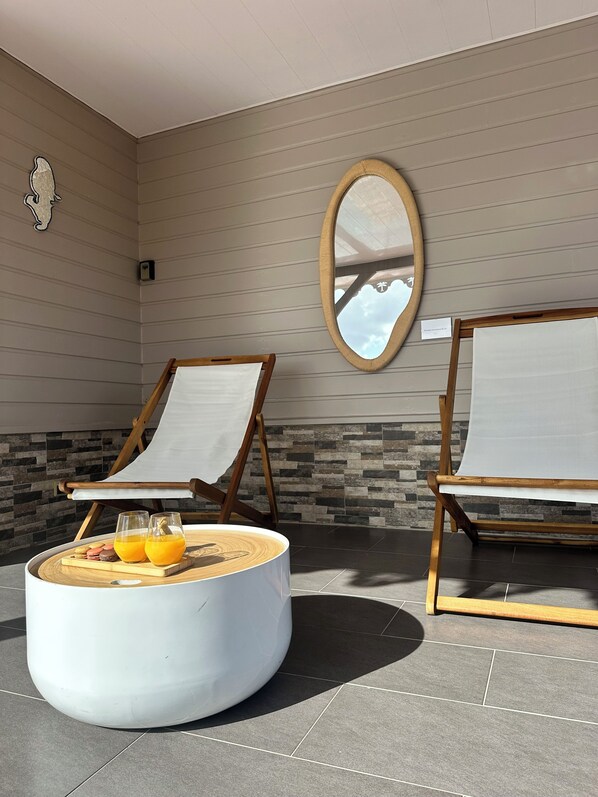 A sunlit room with two lounge chairs, a round coffee table with drinks, and a mirror on the wall.