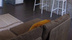 Een moderne woonkamer met een bruine bank, een witte stoel en een geel kussen. Er is een keukenhoek met een witte hanglamp en een roestvrijstalen apparaat.