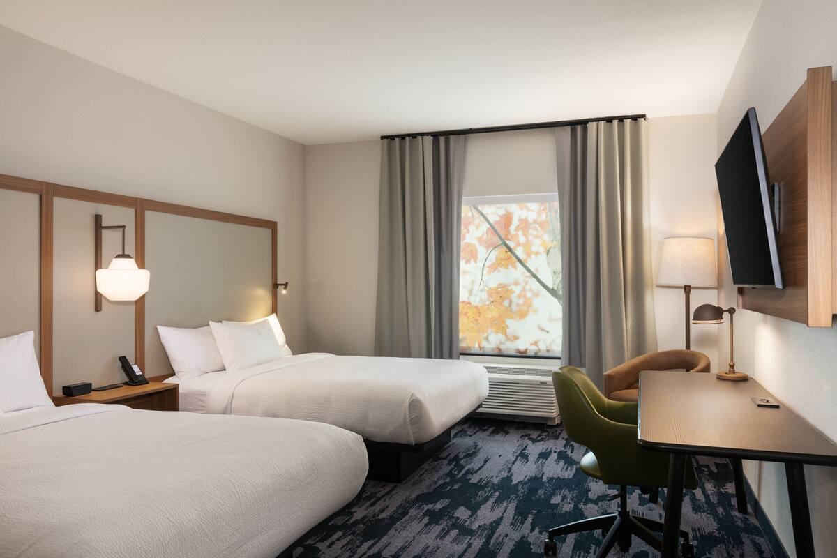 Une chambre d’hôtel avec deux lits, un bureau, une chaise, une télévision, une lampe et une fenêtre avec des rideaux.
