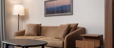A room with a brown sofa, a small round table, a wooden side table, and a framed picture on the wall.