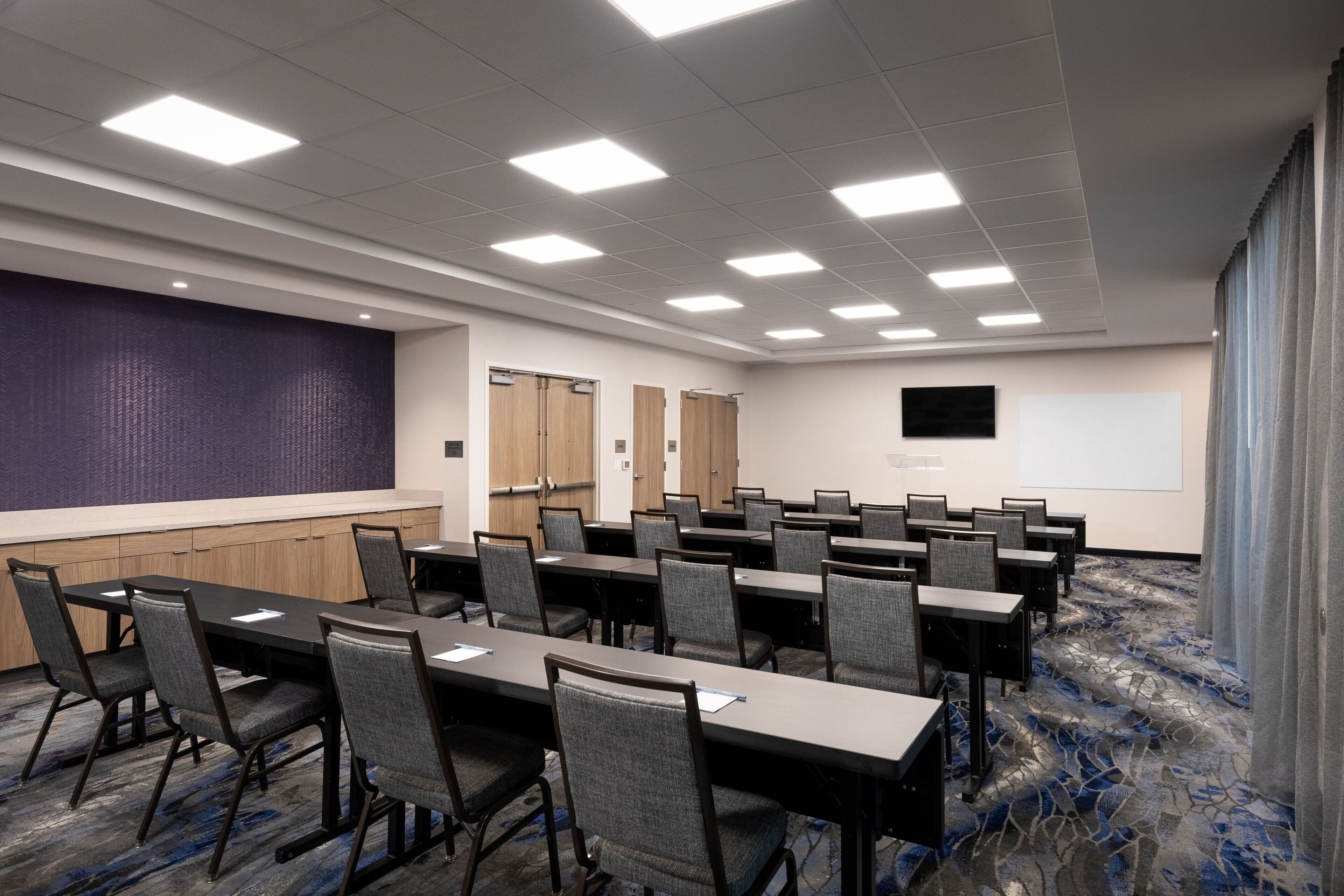 Sala de conferências com várias mesas e cadeiras, uma tela de projetor e uma janela grande com cortinas.