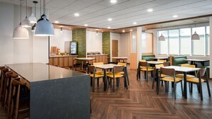 A modern dining area with a long counter, multiple tables, and chairs. There are hanging pendant lights and a kitchen area visible in the background. - Fairfield by Marriott Inn & Suites Omaha at MH Landing (Omaha)