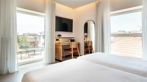 Una habitación de hotel con una cama grande, un escritorio con una silla, un televisor y un espejo. Hay ventanas amplias con cortinas.
