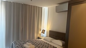 A hotel room with a bed, a bedside table, a window with curtains, and a wall-mounted air conditioner. - Brisas do Lago - Apartamento 4 (Brasília)