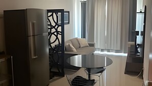 A modern kitchen with a refrigerator, dining table, and chairs. There is a living area with a sofa and a view of the outside. - Brisas do Lago - Apartamento 4 (Brasília)