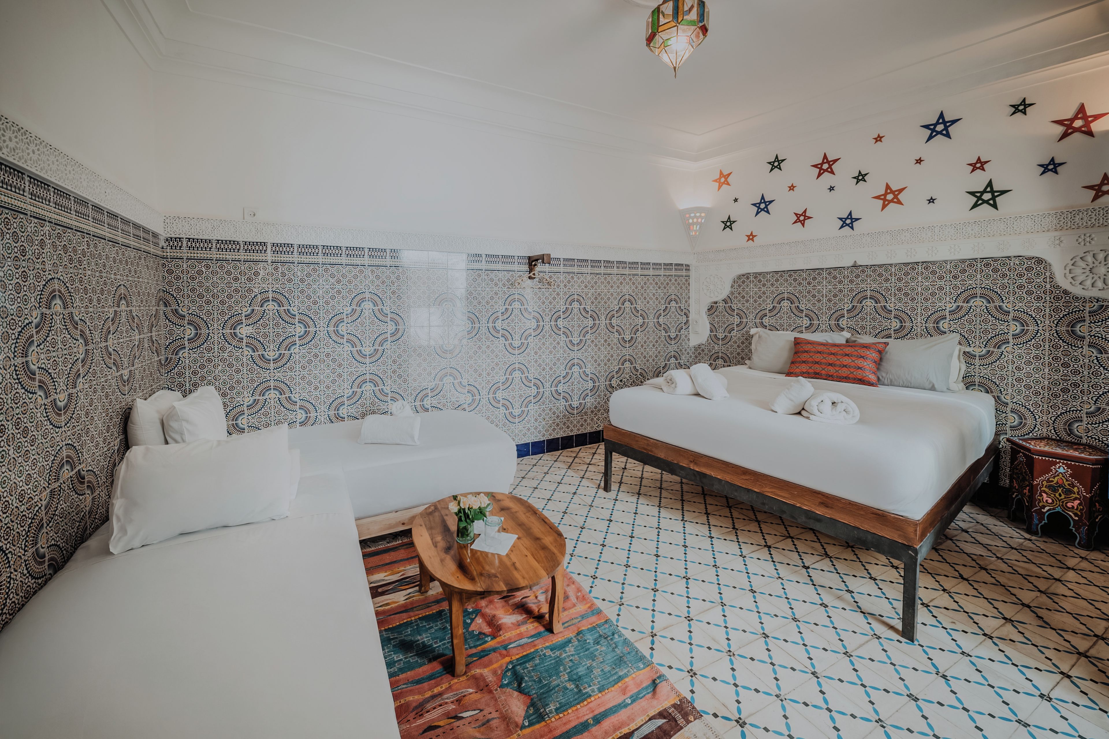 Une chambre d’hôtel avec un mur à motifs, un lit recouvert de draps blancs, une petite table ronde en bois et un tapis coloré.