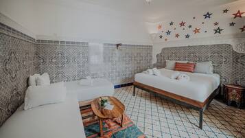 Une chambre d’hôtel avec un mur à motifs, un lit recouvert de draps blancs, une petite table ronde en bois et un tapis coloré.