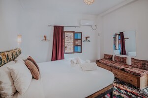 Una habitación de hotel con una cama grande, un banco de madera, un espejo y una ventana con cortinas.