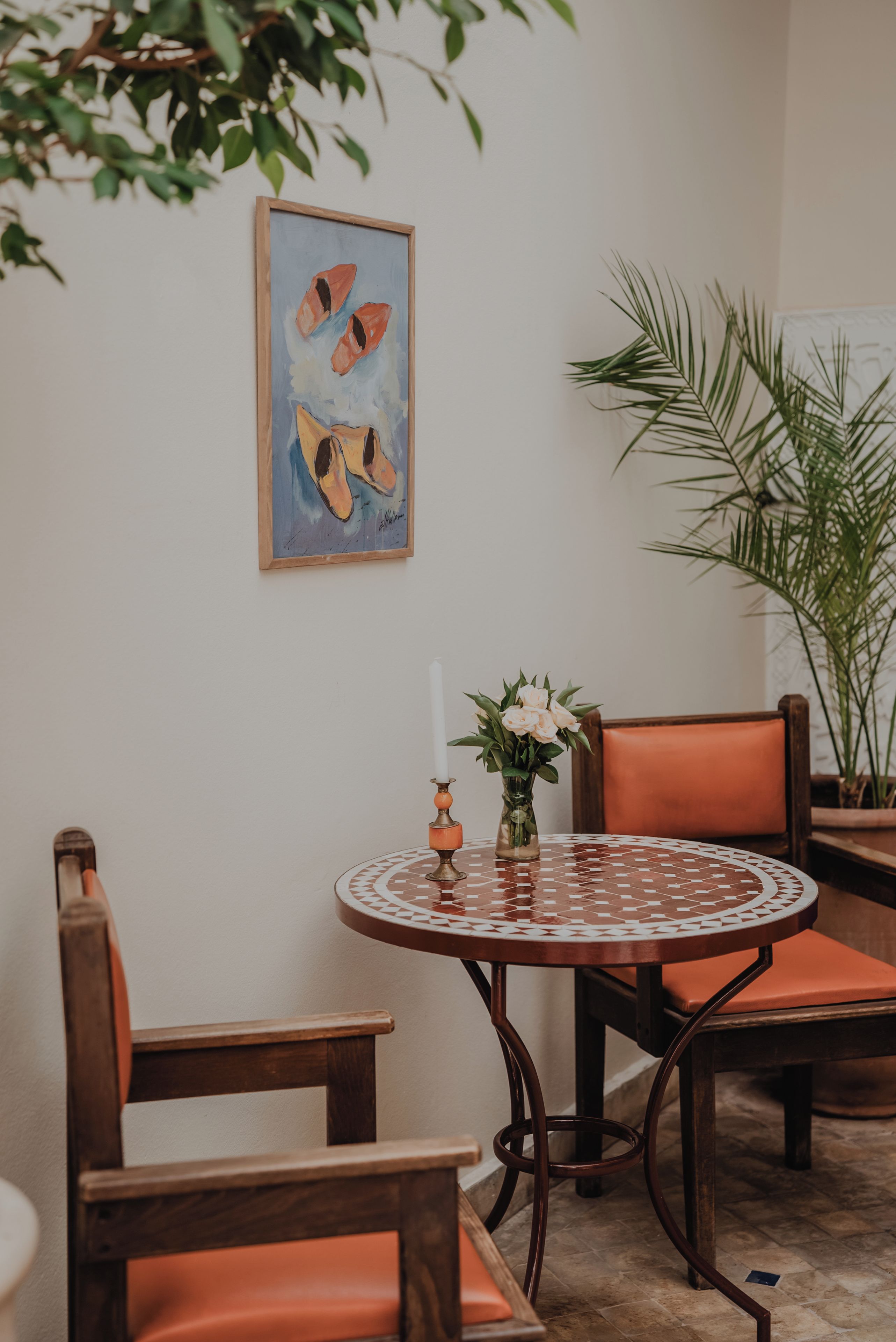 Uma mesa redonda com toalha estampada, rodeada por cadeiras de madeira com almofadas laranja. Um vaso com flores está sobre a mesa.