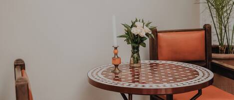 Una mesa redonda con mantel estampado, rodeada de sillas de madera con cojines naranjas. Un jarrón con flores está sobre la mesa.