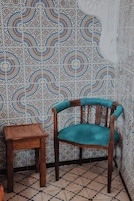 Una habitación con una pared de baldosas estampadas, una silla de madera con cojín y una pequeña mesa de madera.