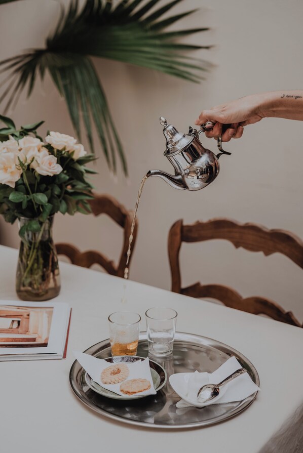 A hand pouring tea from a metal teapot into a glass on a tray with a biscuit and a spoon.